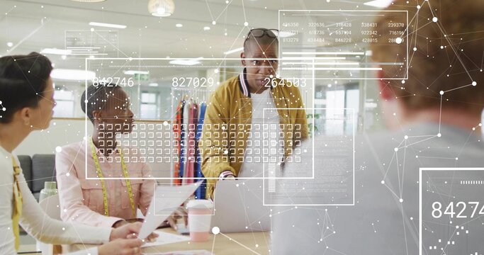 Presenting man in mustard jacket leaning on laptop in office with clothing rack and measuring tape