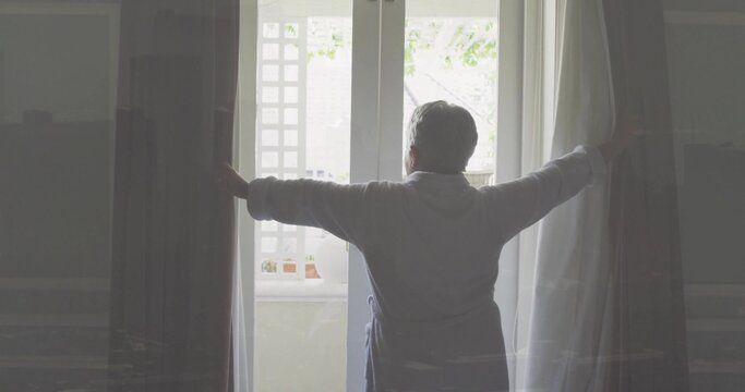 Parting senior woman wearing light robe, standing at divided-pane window and holding heavy curtains - Powered by Adobe