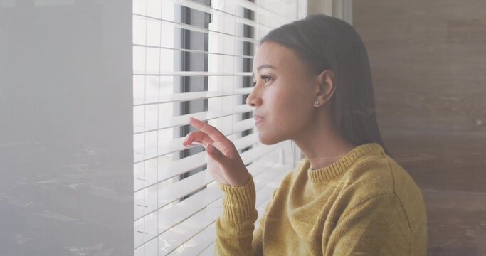 Peering mid-adult woman parting horizontal blinds at window in room, mustard sweater, copy space