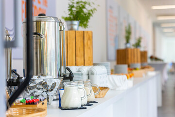 Buffet with a large coffee dispenser, milk jugs, sugar, and tea in the foreground. Cups, plates, and some decorations are visible in the background on a bright table.