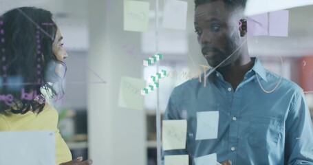 Gesturing coworkers in yellow-top and blue-shirt discussing plan in office, glass with sticky notes