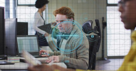 Working redhead in grey sweater typing on laptop in office with hologram security overlay, headset