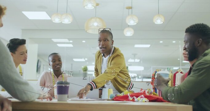 Leaning forward presenter wearing mustard bomber jacket gesturing at wooden table, with red garment