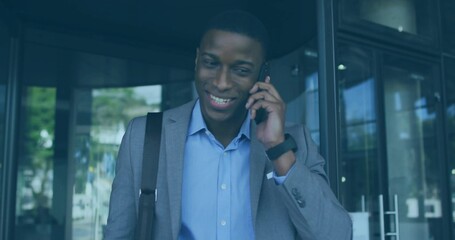 Talking man wearing grey suit and blue shirt exiting glass doors with phone, shoulder bag, watch