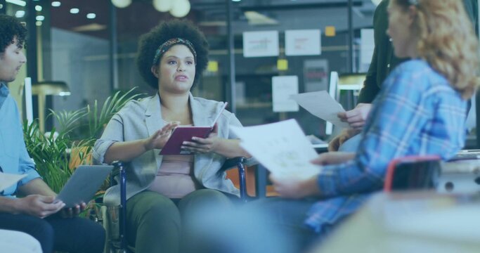 Leading woman in wheelchair in blazer holding tablet, speaking in office with laptop, printouts - Powered by Adobe