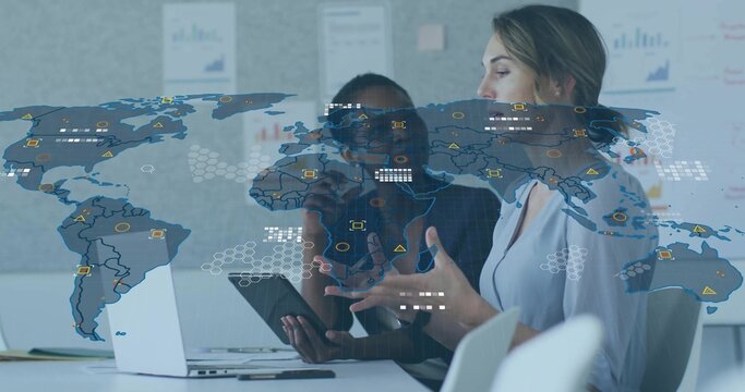 Analyzing two mid-adult women wearing tops, collaborating at meeting table with tablet, map overlay