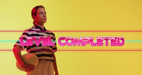 Posing woman in striped jersey, yellow studio, holding ball with LEVEL COMPLETED neon, copy space