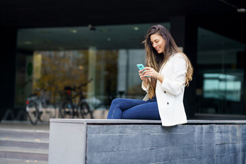 Fototapeta premium A young woman is sitting outdoors, happily texting on her phone while enjoying the day