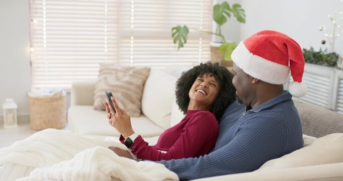 After picking up phone African American couple in Santa hat scrolling photos and smiling on couch