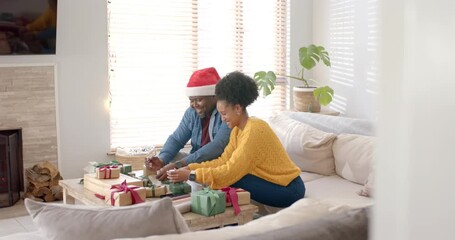 Examining gift tag, diverse couple wrapping holiday gifts at home exchanging smiles - Powered by Adobe
