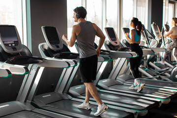 Strong man running on treadmill in well-lit gym, wearing workout clothes and headphones, focused on fitness routine in the morning