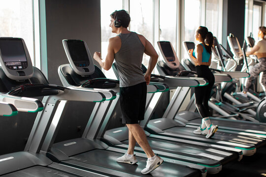Strong man running on treadmill in well-lit gym, wearing workout clothes and headphones, focused on fitness routine in the morning
