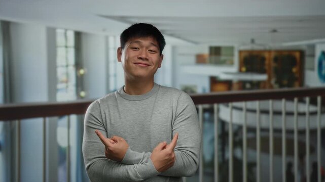 Young chinese man indoors in hotel environment wearing casual grey sweater posing playfully pointing fingers in opposite directions highlighting modern interior background.