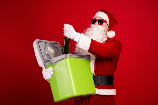 Santa in red costume with beard and glasses holds a bottle over a bright green cooler against a solid red background for a festive stock photo listing