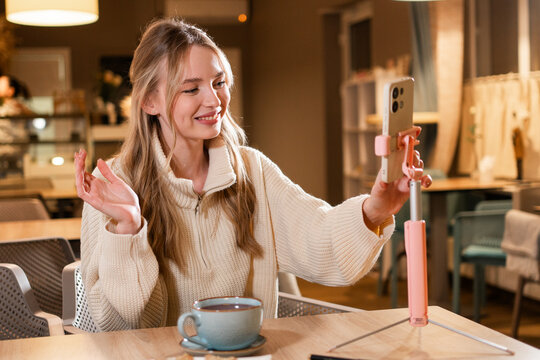Young smiling woman having video call or chatting using mobile phone. freelancer girl waving hand on smartphone during remote work in cozy cafe