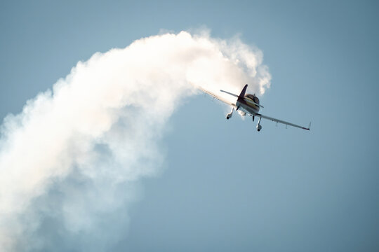 Small aerobatic aircraft flying upward leaving a thick white smoke trail during an air show against a bright blue sky.