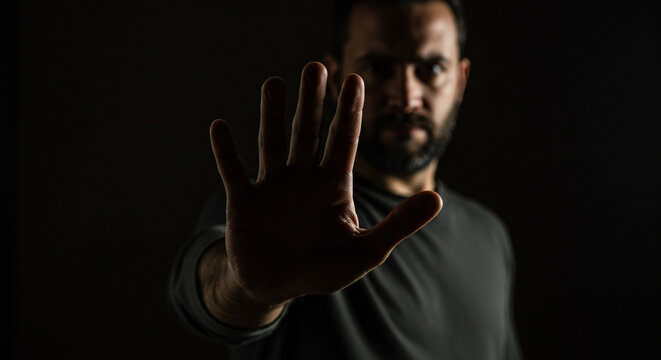 Intense man with beard holds up hand in stop gesture against dark background conveying warning or disagreement - Powered by Adobe