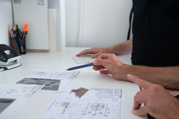 Zwei Personen besprechen technische Zeichnungen und Pl&auml;ne an einem wei&szlig;en Schreibtisch, Fokus auf Hand mit Kugelschreiber und die Dokumente, moderne B&uuml;roatmosph&auml;re.