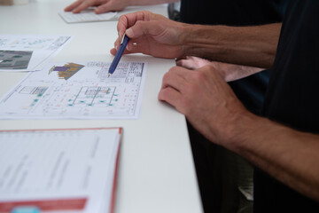 Zwei Personen besprechen technische Zeichnungen und Pl&auml;ne an einem wei&szlig;en Schreibtisch, Fokus auf Hand mit Kugelschreiber und die Dokumente, moderne B&uuml;roatmosph&auml;re.