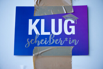 Lustiges Schild &bdquo;Klugschei&szlig;er*in&ldquo; mit Klebeband an Wand befestigt