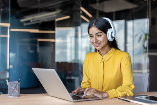 Smiling Indian young woman wearing headphones sitting in office and working on laptop