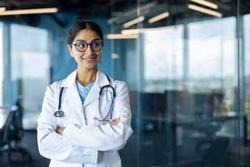 Young Indian female doctor, intern in uniform standing in a hospital room, arms crossed on chest and looking to the side with a smile