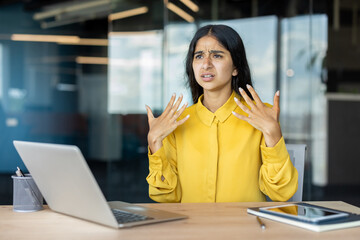 Young Indian woman sitting in office at desk, waving hands in face, suffering from heat and suffocation