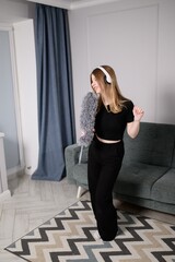 Caucasian woman dances in living room with headphones, listening to music while cleaning and singing into pipidaster. Expressing happiness through dance and music. Concept of order and cleaning.