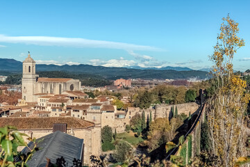 Panoramic view of Girona’s old town with the Cathedral of Saint Mary rising above medieval buildings and ancient walls, framed by snow-capped Pyrenees mountains under a clear blue sky, Spain