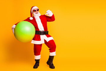 Santa in red costume holding large green ball smiles against yellow background festive cheerful Christmas holiday promotion and family themed imagery