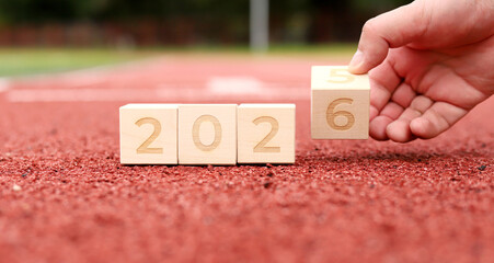 A hand places a wooden block showing the number six next to blocks showing 2026, symbolizing the transition to the new year. A joyful atmosphere celebrates the upcoming year