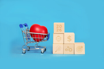 A small shopping cart holds a bright red heart, representing love and hope. Adjacent to it are wooden blocks with symbols for growth and money, celebrating New Year's aspirations for 2026