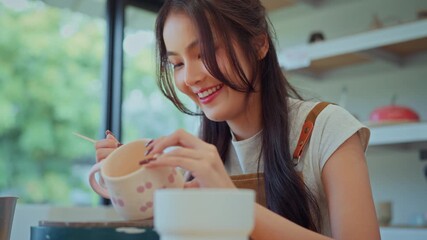 Asian woman smiling while painting a ceramic mug by hand, focusing on adding decorative patterns during a pottery workshop, enjoying sculpting process and creative small business lifestyle