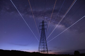 Long Exposure View Of High-Voltage Electricity Transmission Tower Against A Clear Deep Blue Night Sky With Trailing Lights From The Power Lines And Distant Stars
