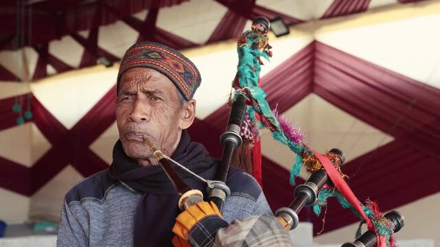 4K footage of an elderly Uttarakhand village musician playing a traditional bagpipe during a marriage celebration. Close-up view capturing his expressions, cultural attire, and festive atmosphere.
