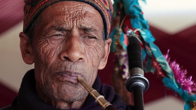 4K footage of an elderly Uttarakhand village musician playing a traditional bagpipe during a marriage celebration. Close-up view capturing his expressions, cultural attire, and festive atmosphere.
