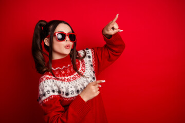Young woman in red knit sweater with Nordic pattern and sunglass poses playfully against a red background in a festive Christmas fashion look