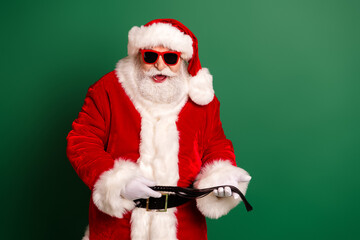 Santa in red suit with white fur trim wearing glassed and smiling while holding a belt against a green background for Christmas promotion