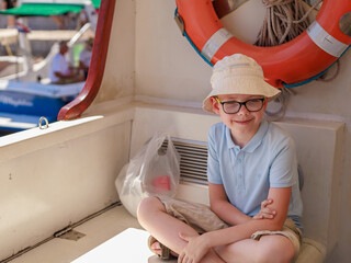 Smiling boy in a hat sitting on a boat deck near an orange lifebuoy on a sunny day. Concept of family travel, summer adventure and positive outdoor lifestyle.