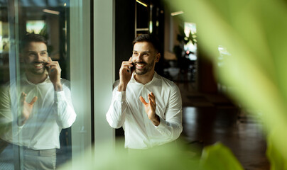Smiling man talking on the phone near a large window in a modern office setting