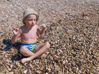 Child sitting cross-legged on a pebble beach with closed eyes and wide copy space around. Concept of relaxation, childhood mindfulness and natural outdoor lifestyle.