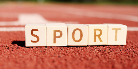 Wooden blocks spell the word sport, arranged on a red running track under bright sunshine. The setting creates a vibrant and energetic atmosphere for athletic activities