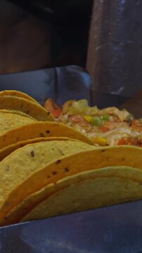 Vertical video. Row of Hard-Shell Tacos Standing Upright beside Cooked Chicken and Vegetables on Grill Station, Concept of Mexican Street Food and Culinary Tradition