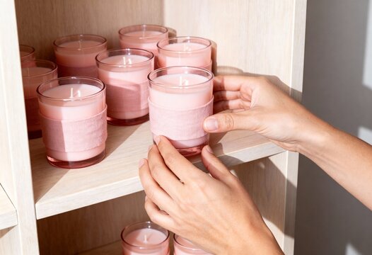 Woman's hands placing a handmade pink candle on a wooden shelf. Small business owner organizing aromatherapy products. Home decor and wellness concept