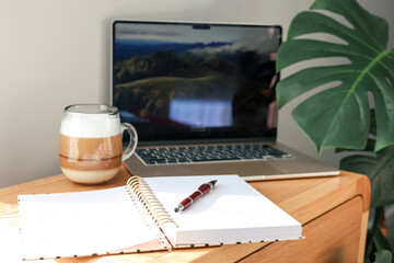 A bright and cozy home workspace with a laptop, notebook, pen, and a cup of layered coffee on a...