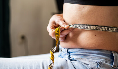Close up of faceless woman in jeans measuring her waist with yellow tape measure. Weight loss