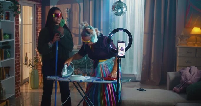 Grandmother speaking to the audience while looking at a smartphone mounted on a ring light, pretending to DJ with her granddaughter as they play music, dance and create fun social-media content at hom