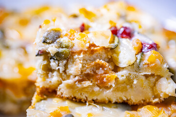 A thick slice of fruit and rhubarb cake topped with pumpkin seeds, dried cranberries and apricots, removed from a round baked cake. Rich texture and visible moist layers.