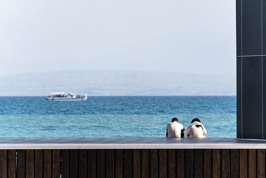 A couple sits on a wooden pier overlooking a vast blue sea or lake on a sunny day. In the distance, a small tourist boat is visible, and hazy mountains line the horizon. - Powered by Adobe