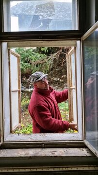 Senior man installing an old wooden window into an abandoned rural house frame during DIY restoration work